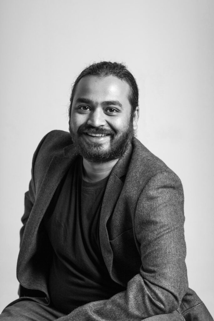Black-and-white professional portrait of a man with a beard and tied-back hair, dressed in a blazer and dark t-shirt, smiling warmly while seated against a plain studio background.