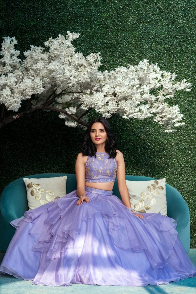 Since I don't have the image, I will base the descriptions on a full shot of the model sitting on a couch, still wearing the lavender lehenga, allowing the skirt to fan out beautifully, likely in an indoor studio or luxurious living room setting. Alt Text (Accessibility & SEO) Alt Text: Full-length fashion portrait of a woman sitting on a low couch in a lavender tulle lehenga, showcasing the wide volume of the skirt and the gold-embroidered crop top.