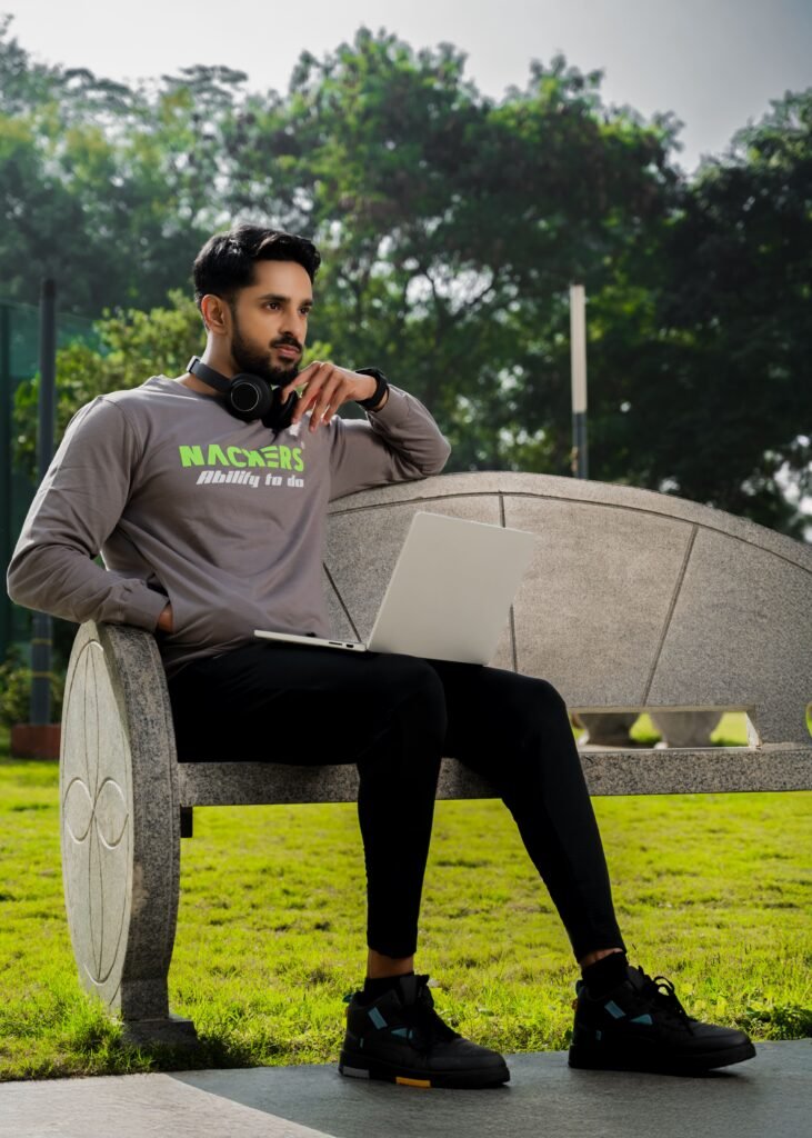 Male model wearing a grey NACKERS long-sleeve graphic tee and black athletic pants, sitting on a stone bench outdoors while working on a silver laptop, with headphones around his neck.