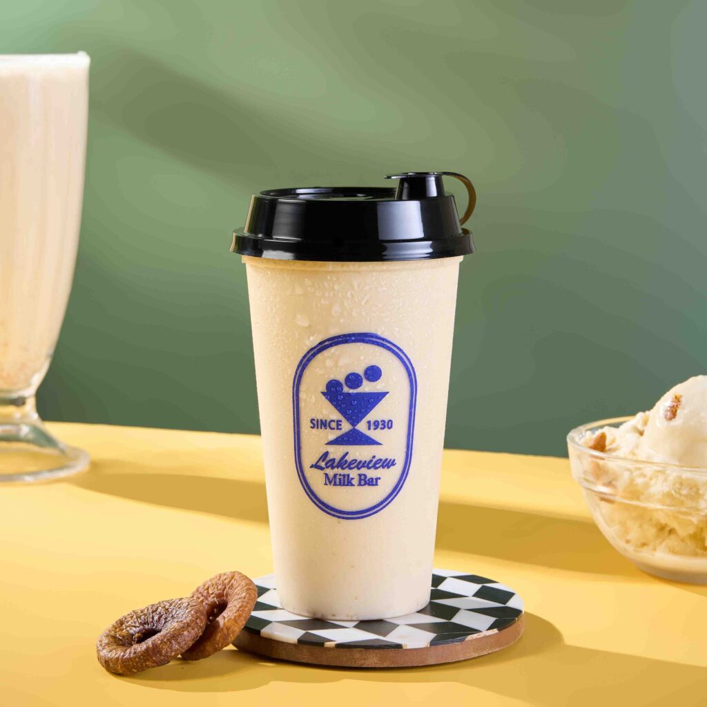 Takeaway cup of a light-colored milkshake or smoothie from Lakeview Milk Bar (Since 1930), resting on a checkered coaster with a small churro and a bowl of ice cream in the background.