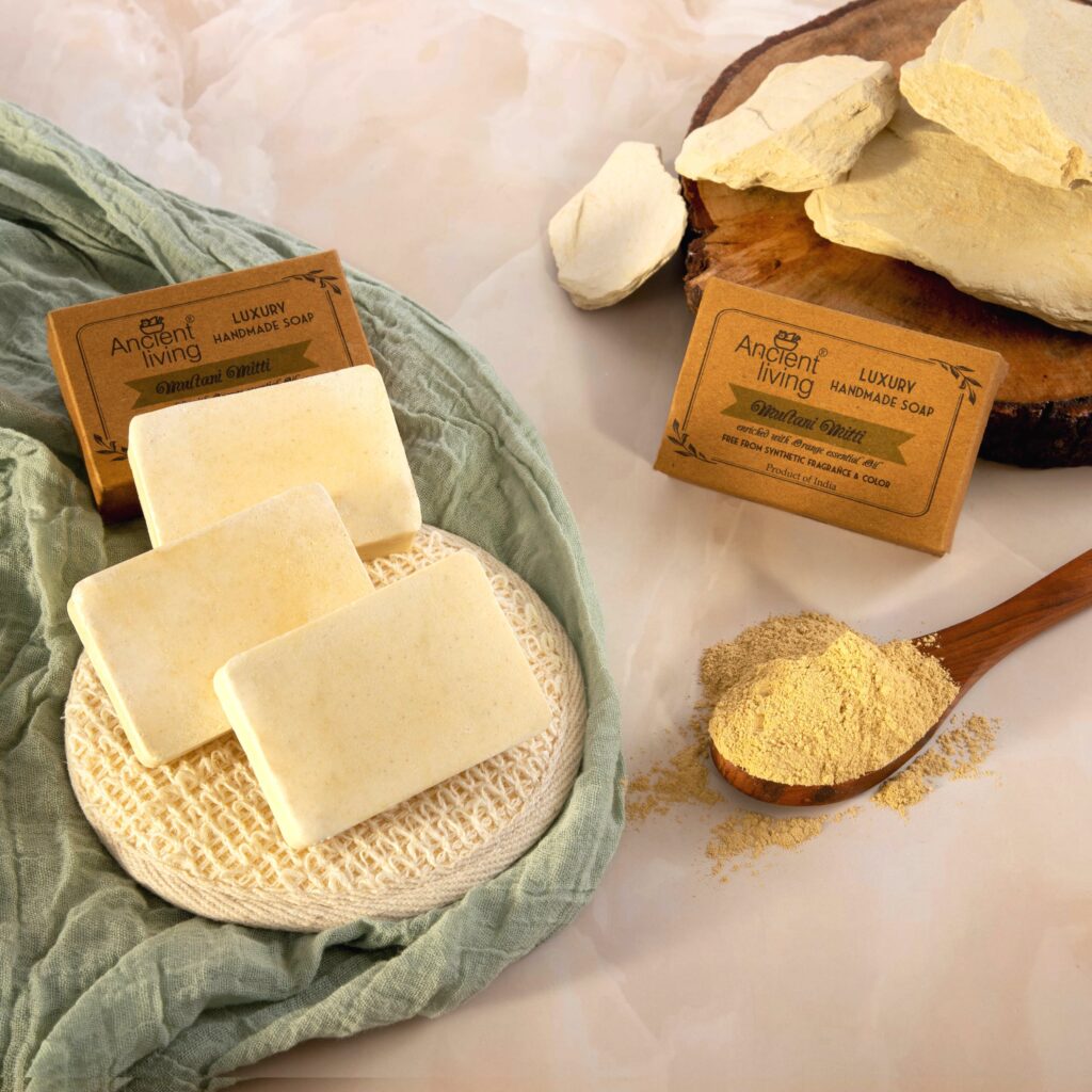 Three rectangular Multani Mitti soap bars displayed with natural clay powder, raw clay chunks, and brown "Ancient Living" product packaging.