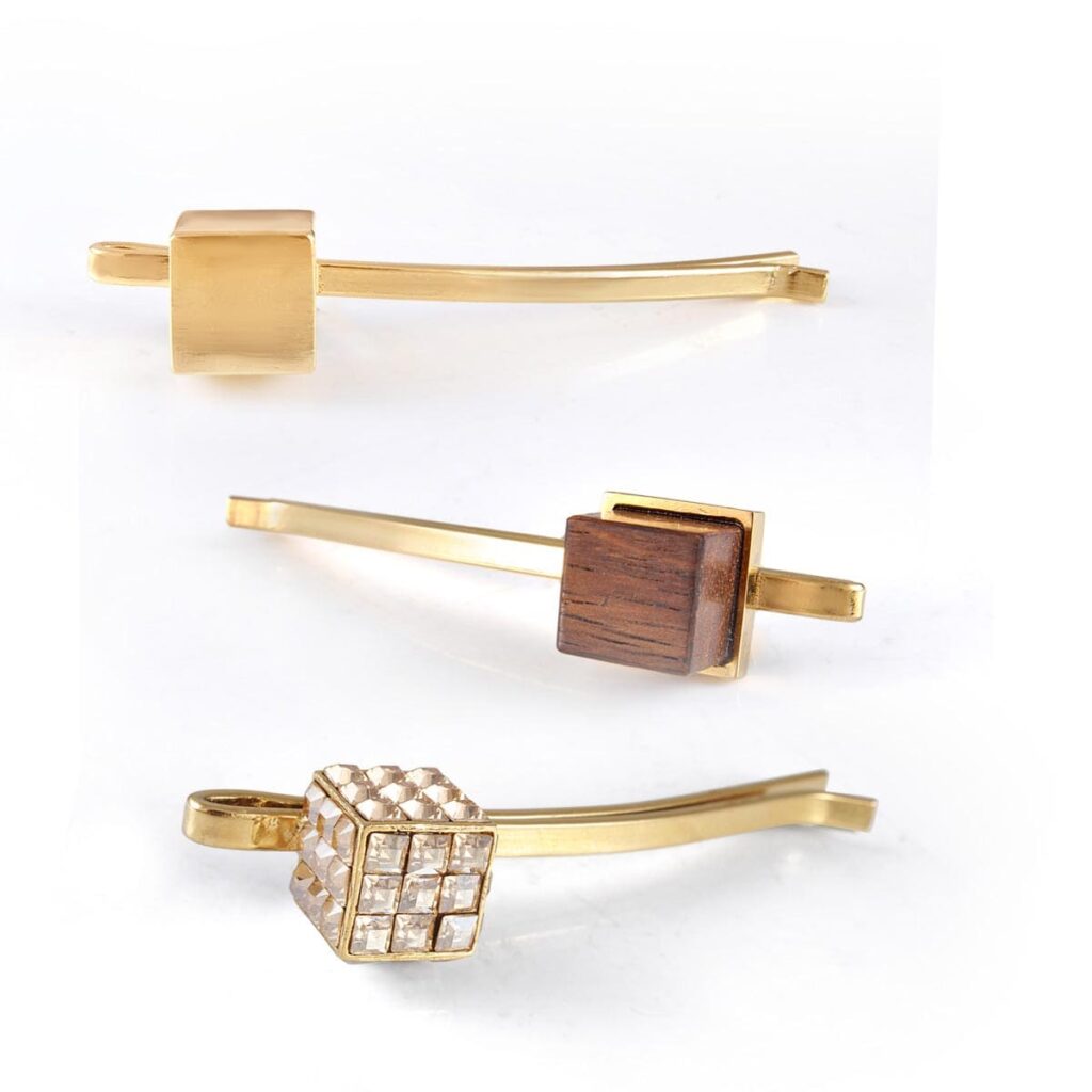 Three modern gold hair clips or barrettes, featuring three different cuboid accents: solid gold, dark wood, and clear pave-set diamonds, arranged horizontally on a white background.