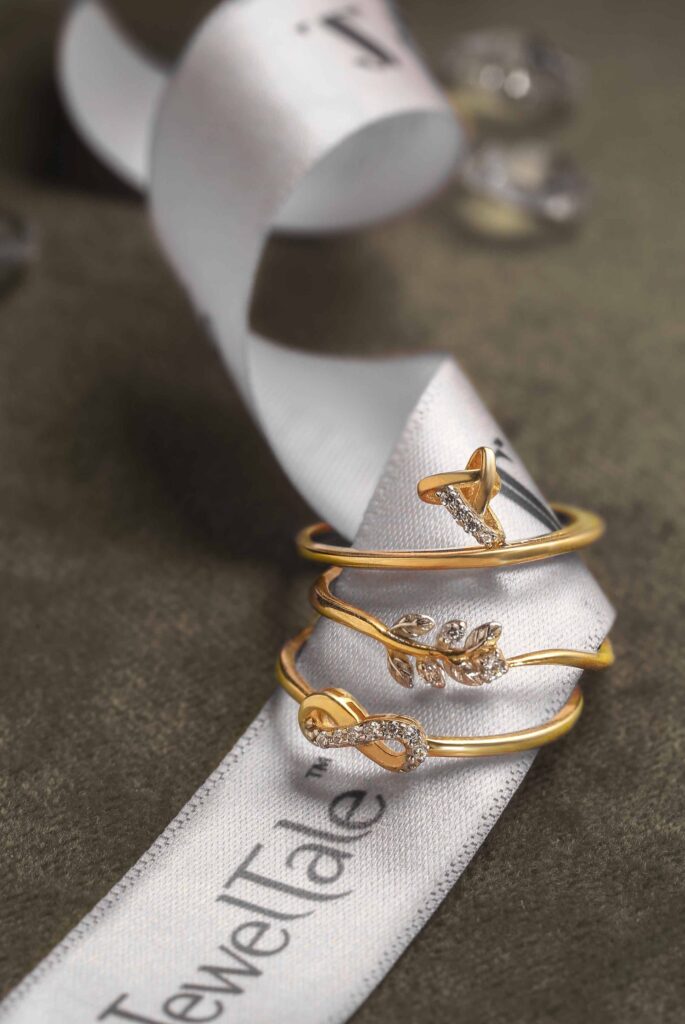 Three delicate gold stackable rings with small diamond accents in butterfly, leaf, and bow designs, resting on a white, branded Jewel Tale satin ribbon against a dark surface.