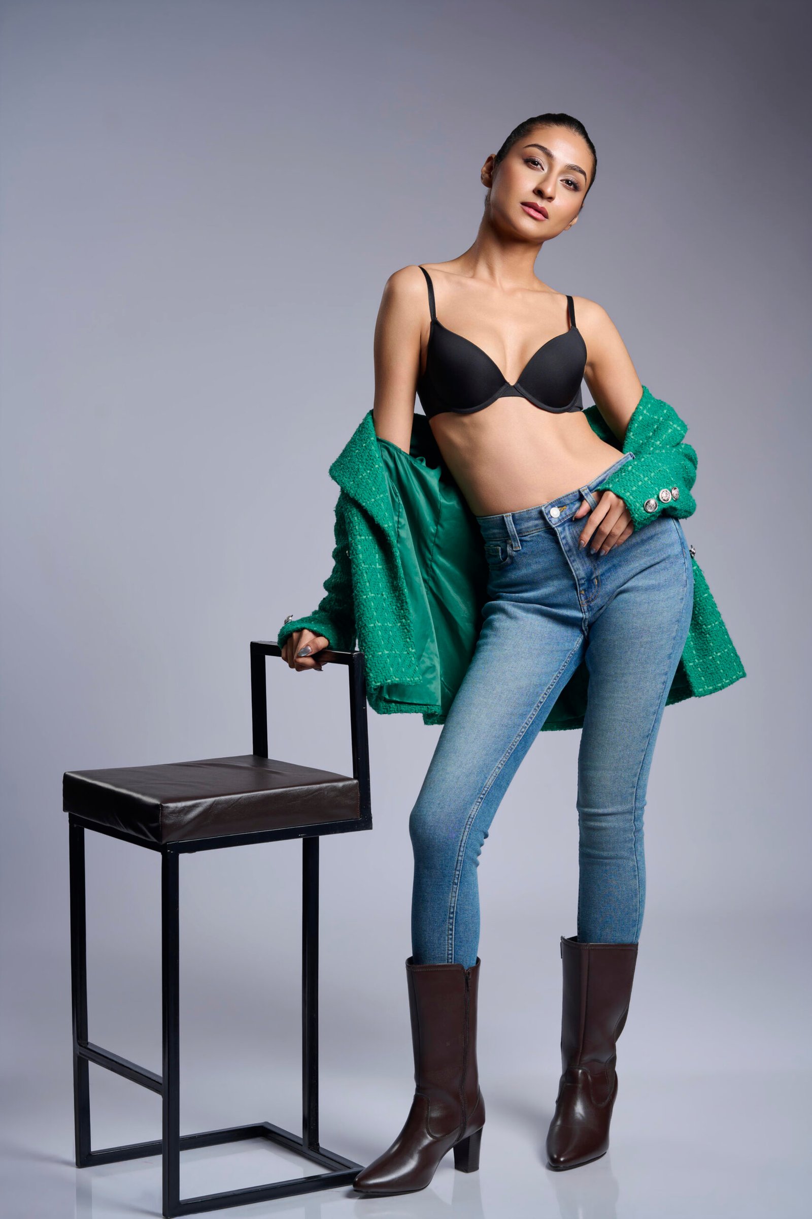 tanding model leaning against a modern dark stool, wearing a black bra, blue denim jeans, tall dark boots, and an open green jacket.