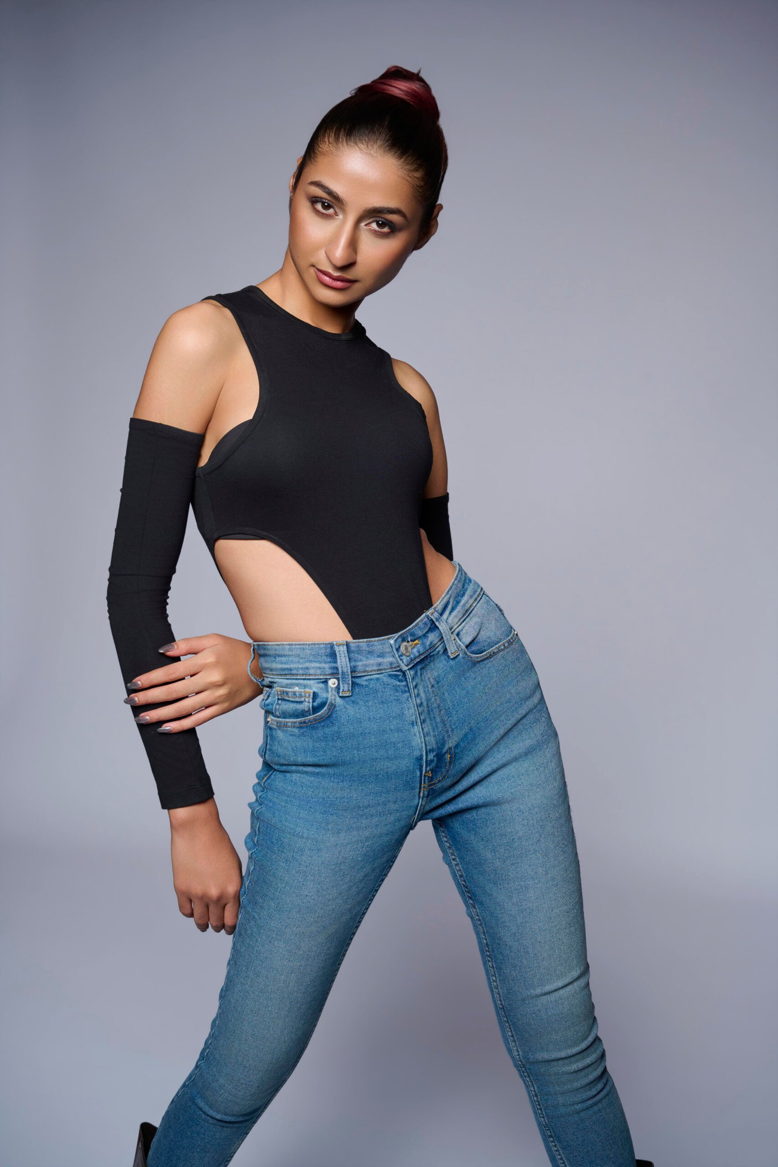 Alt Text: Studio portrait of a model leaning slightly with a confident look, wearing a black cut-out top with detached sleeves and blue denim jeans against a neutral background.